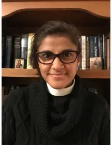 A woman with black hair, brown skin, and glasses, wearing a clerical collar, in front of a bookshelf