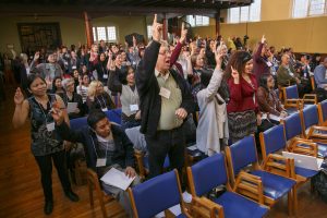 People seated and standing in a large hall raise their arms in the air.