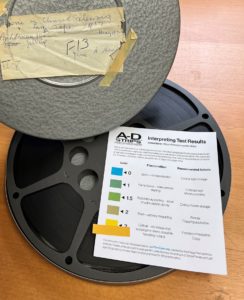 Image showing lid of film canister with title of film, along with 16 mm film reel and bright yellow A-D test strip and result key showing critical deterioration.